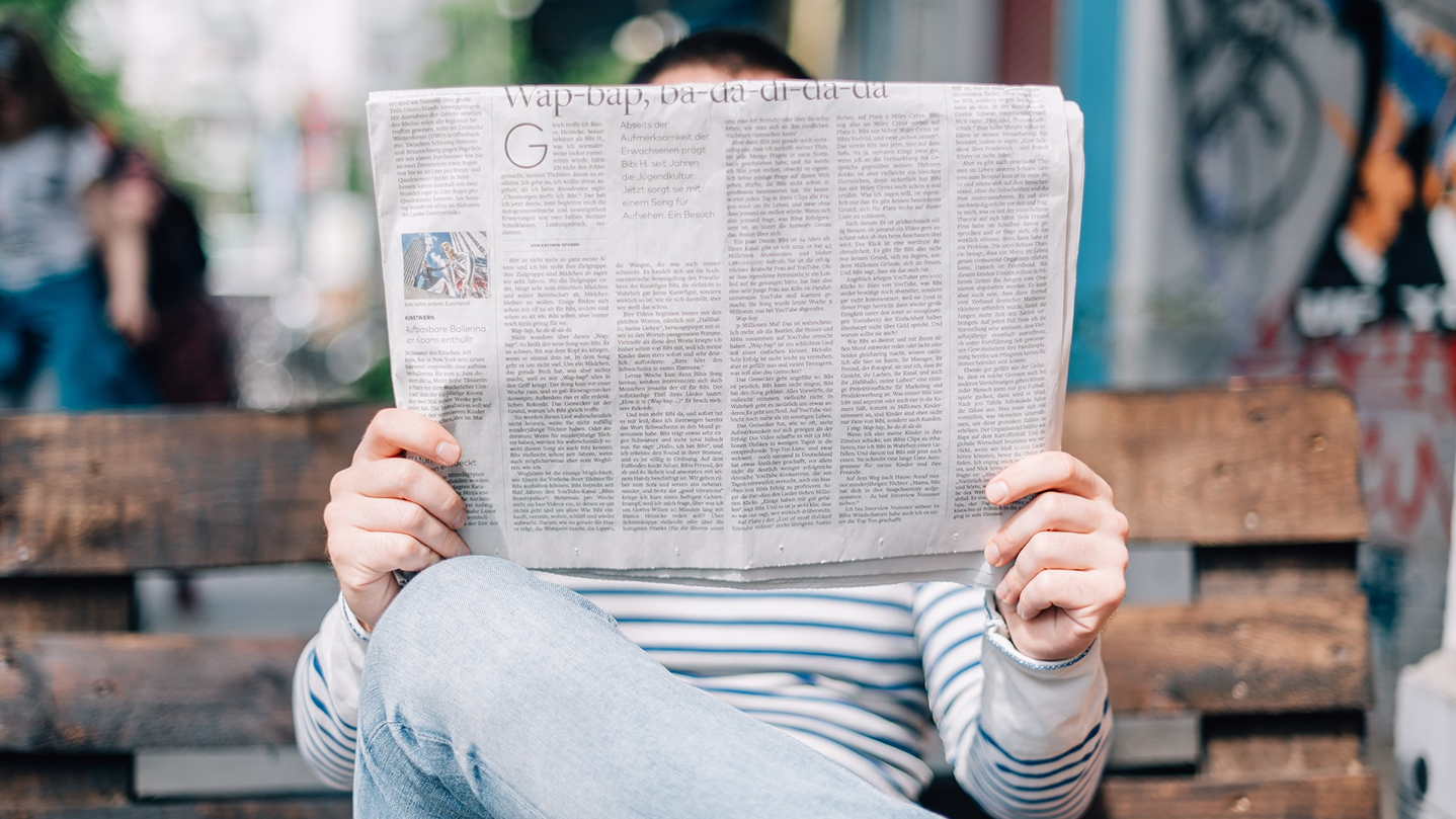 Someone seated on a bench outside, in front of a mural, with their face hidden by the newspaper they're reading.