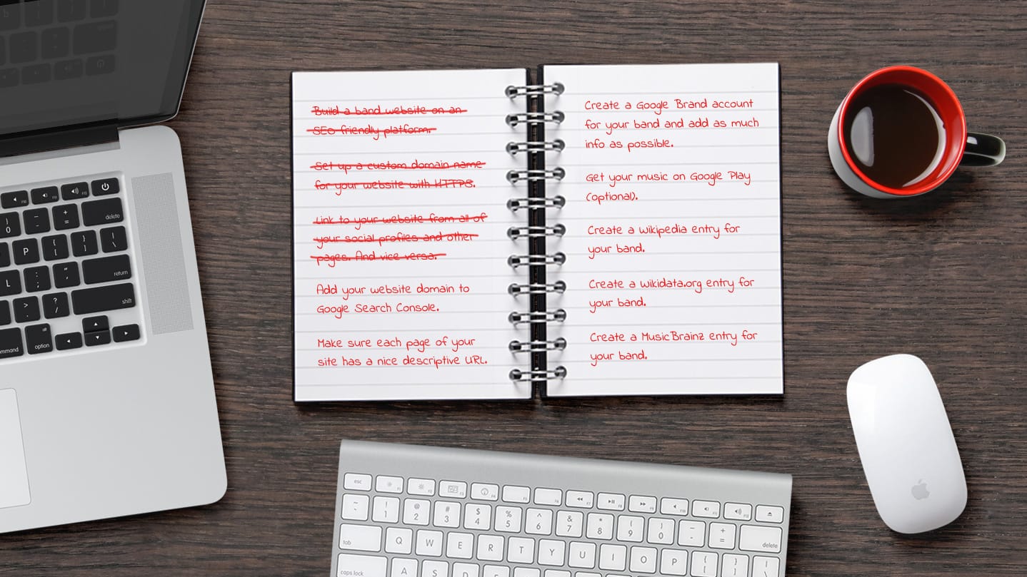 Overview of notebook, keyboard, mouse, laptop, and cup, on a table top.