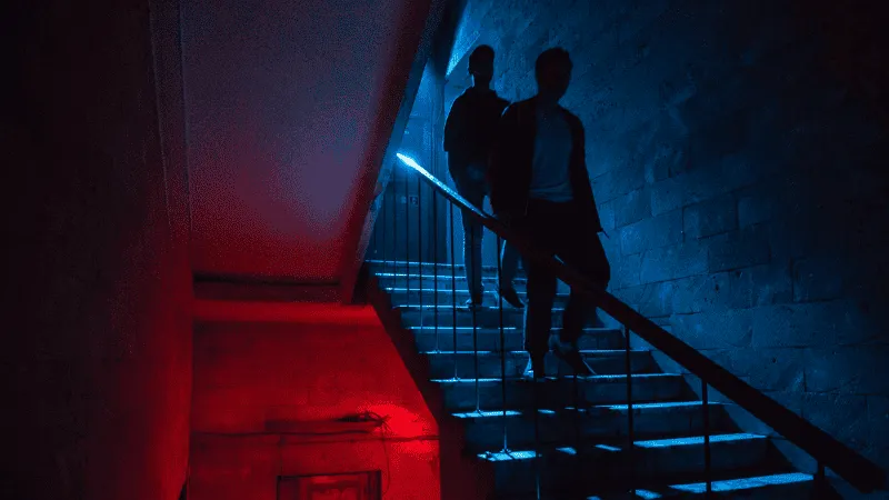Dark urban staircase illuminated with contrasting red and blue lights, featuring two silhouetted figures walking down, creati