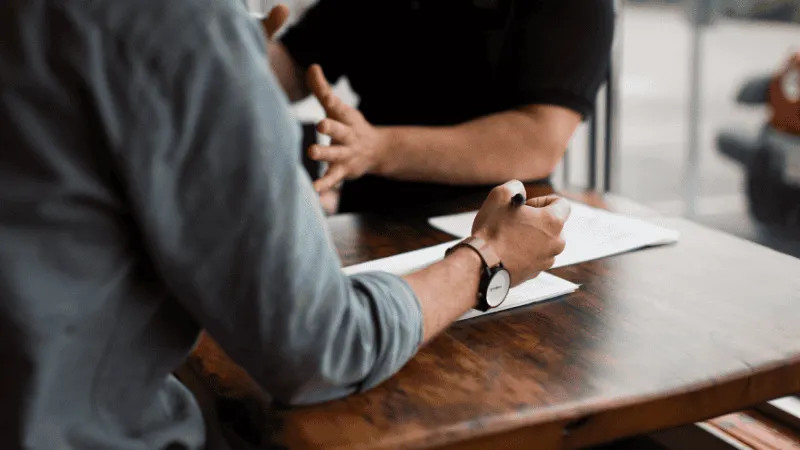 A person signing a document at a wooden table during a business meeting or professional discussion.