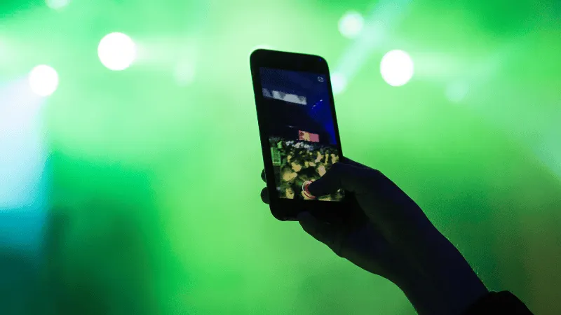 A person holding a smartphone capturing or streaming live music concert with bright green stage lighting in the background.