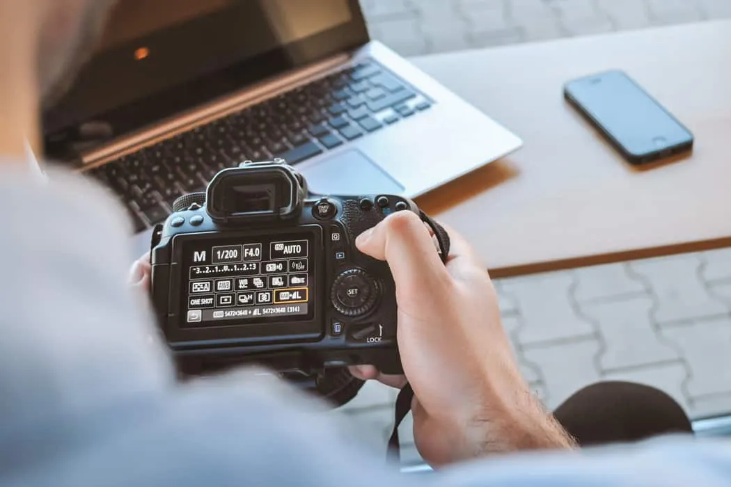 High-quality DSLR camera on photographer's hand with laptop and smartphone on wooden desk, focusing on digital media producti