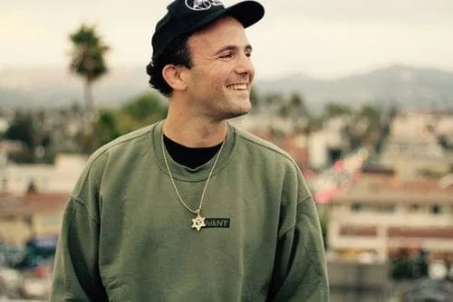 Bright smiling man in casual hoodie and cap outdoors with cityscape background, representing music industry or artist lifesty