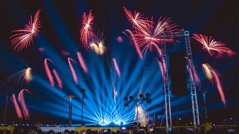 Spectacular fireworks display over a music festival stage with vibrant blue lights and large speaker setup, creating an energ