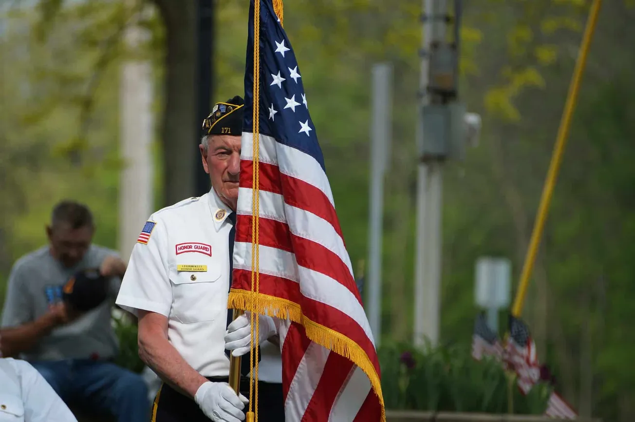Veteran honor guard member holding the American flag during Memorial Day ceremony in a park with trees in the background.