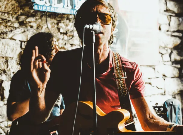 A musician performing live at a venue with stone walls, wearing sunglasses and playing an acoustic guitar and keyboard.