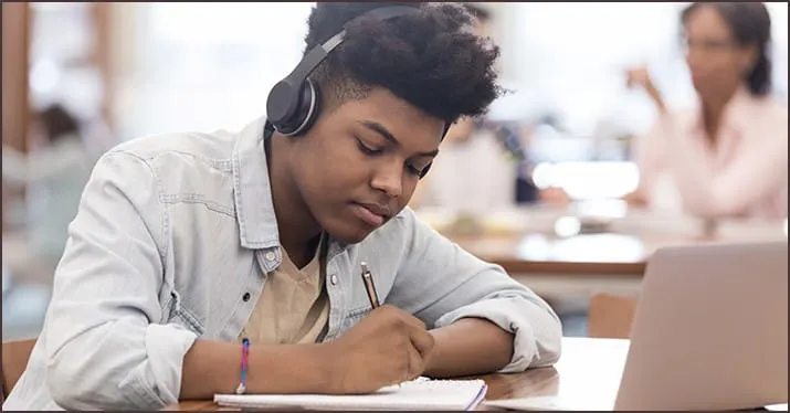 Young student wearing headphones, taking notes on a laptop in a classroom setting for e-learning, digital education, and onli