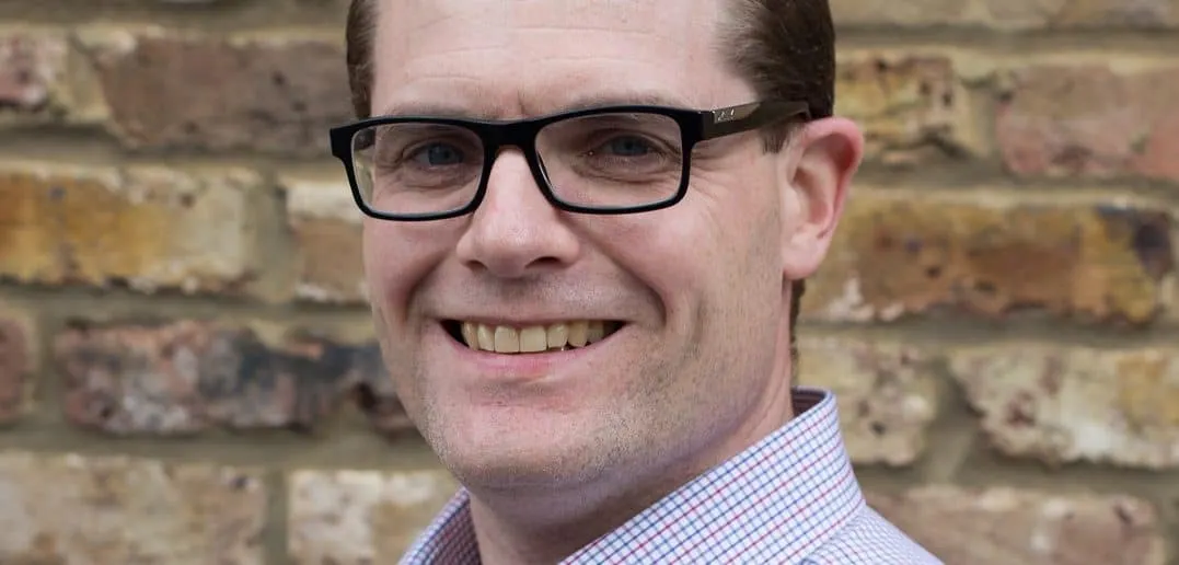 Professional man with glasses smiling in front of a brick wall, representing industry professional, confident speaker, or bus