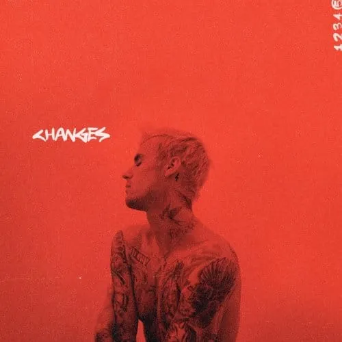 A young tattooed man with platinum blond hair stands against a vibrant red background, creating a striking, modern portrait w