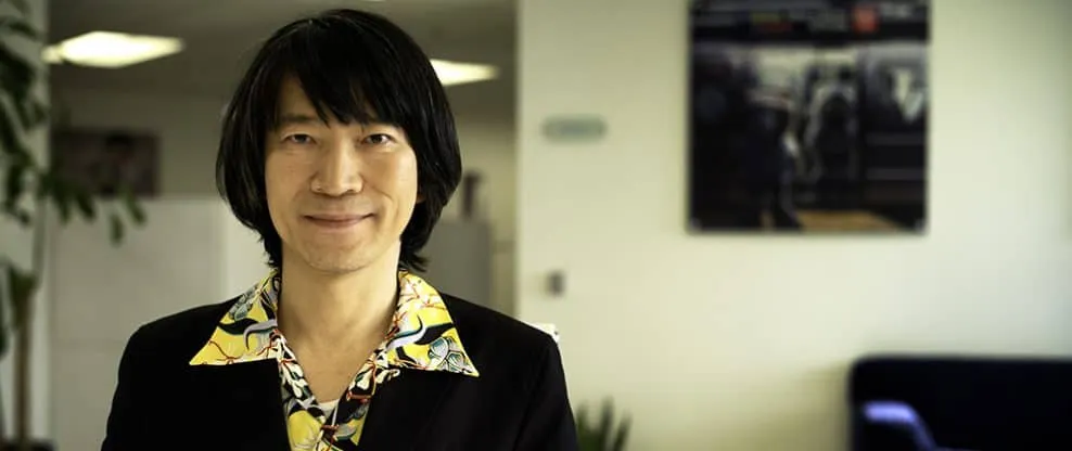 Elegant Asian man in business casual attire smiling in modern office environment with a large TV screen and green plant in th