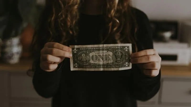 One dollar bill held by a person in a black long-sleeve shirt, small home interior background, symbolizing finance or money c