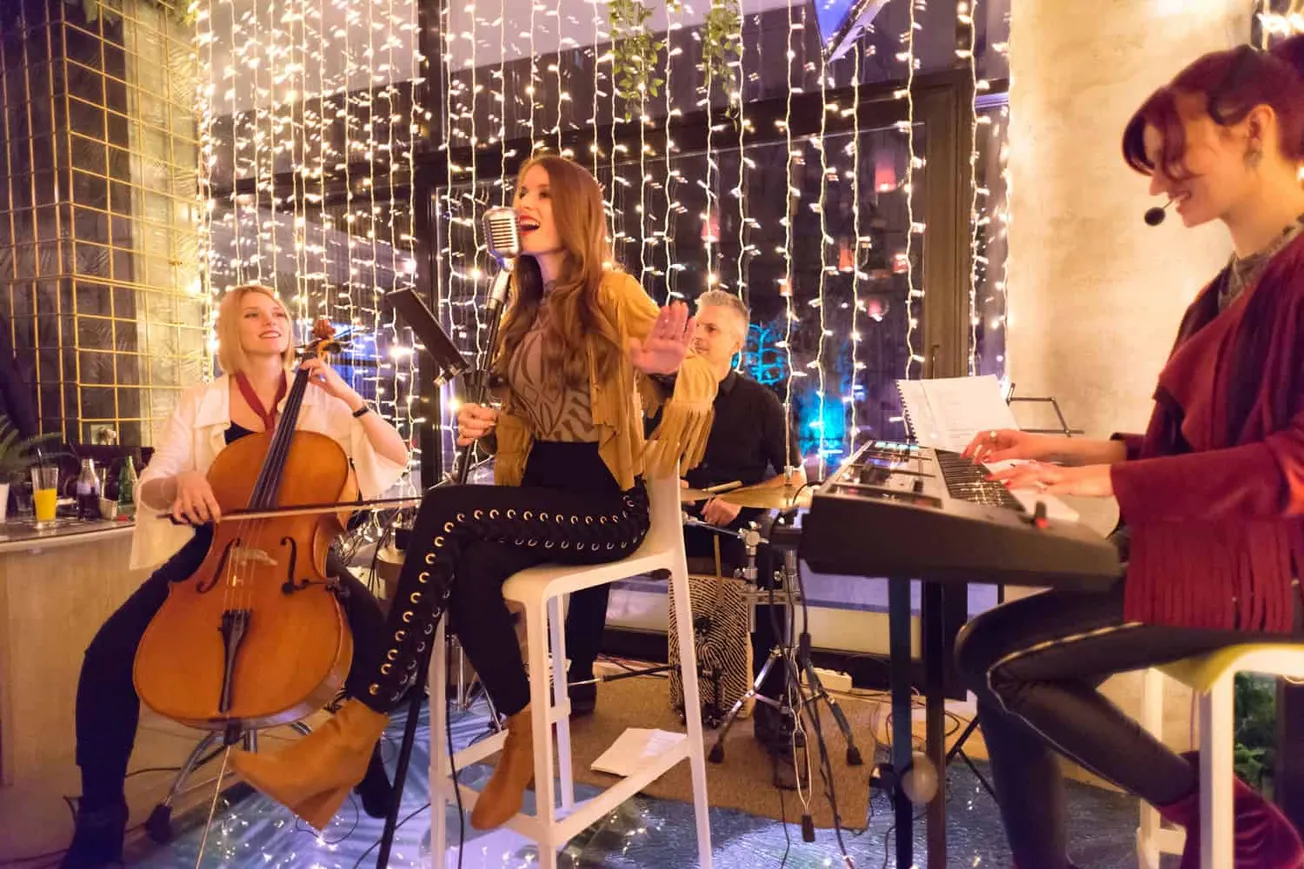 Elegant live music performance featuring a singer, cellist, and keyboardist in a cozy venue decorated with string lights, cap