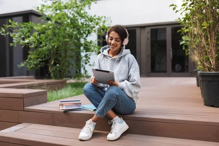 Relaxed young woman sitting outdoors on wooden steps, using a tablet with headphones, studying or working, surrounded by gree