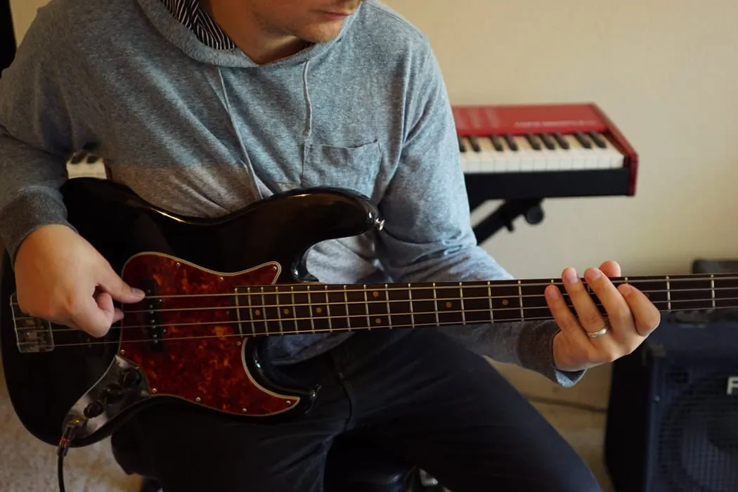 Electric bass guitar playing musician in home music studio with keyboard and amplifier in background.