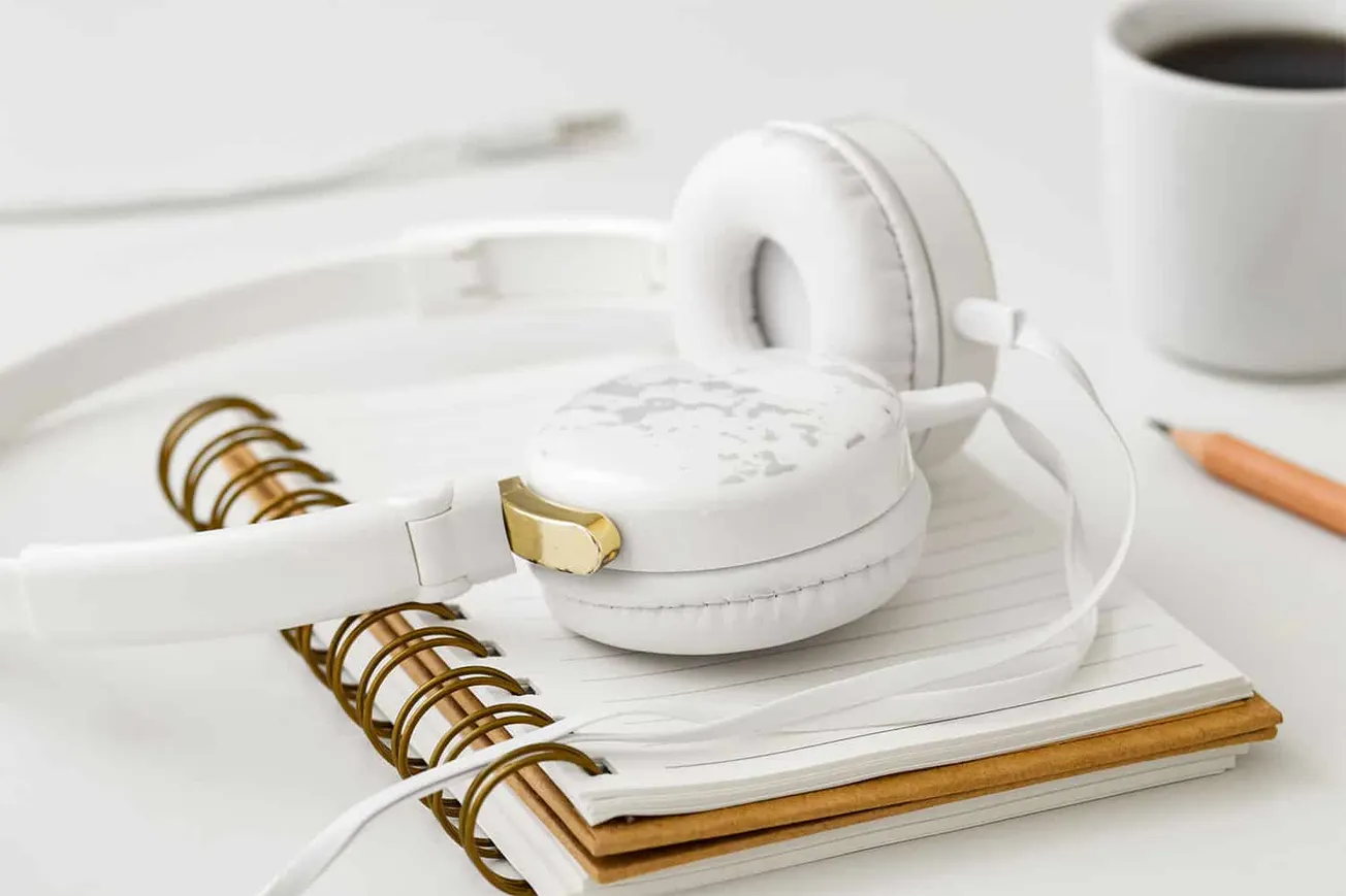 White over-ear headphones resting on a notebook with a gold paperclip, a cup of coffee in the background, and a pencil on a c