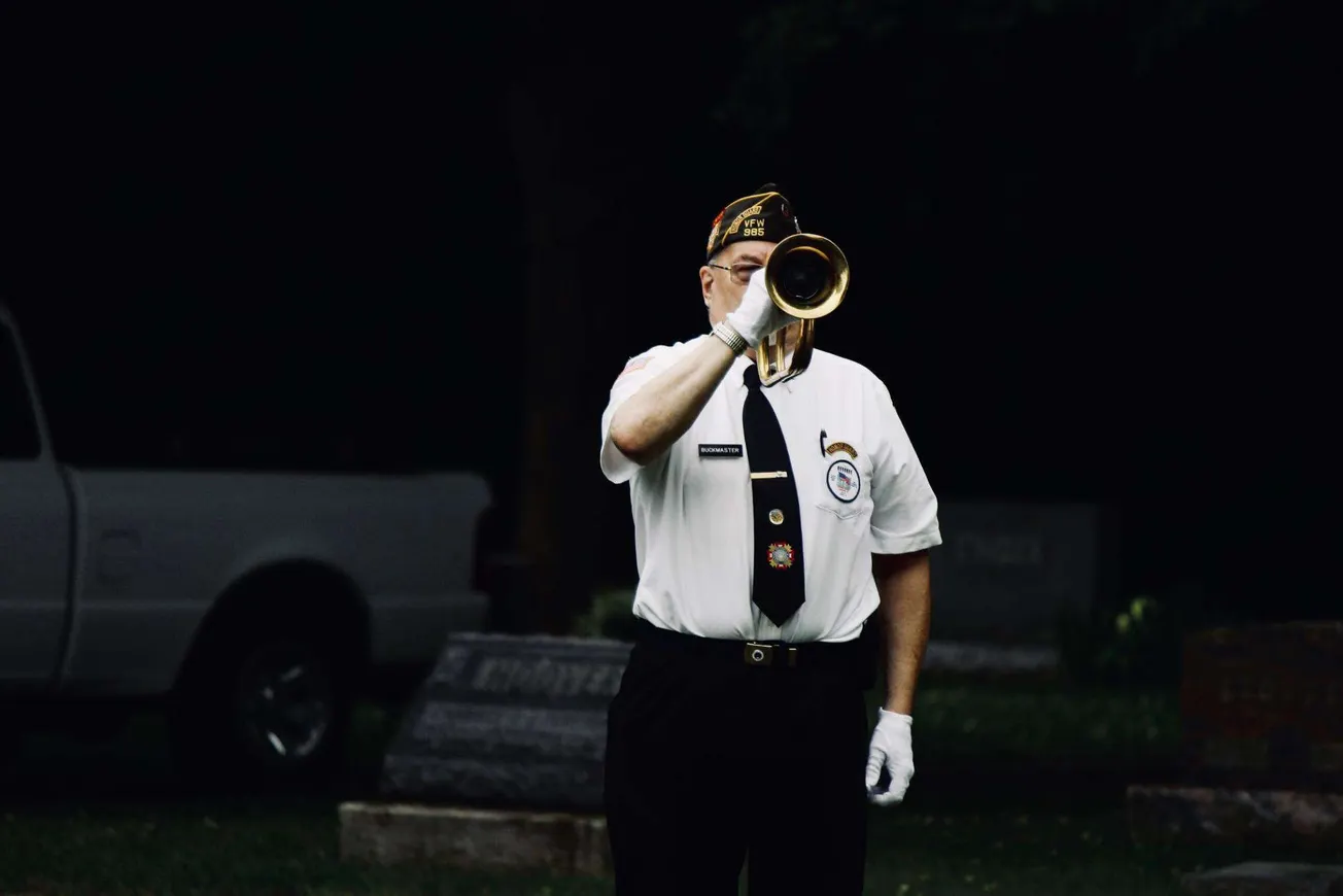 Patriot playing bugle at a memorial ceremony honoring military veterans, wearing ceremonial uniform with badges and patriotic