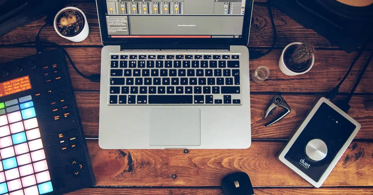 Laptop with music production software on wooden desk, surrounded by audio equipment, coffee cups, and accessories, representi