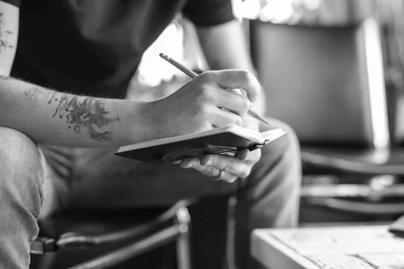 A person writing in a small notebook with a pen, sitting indoors, with a focus on their hand and tattoo; black and white imag