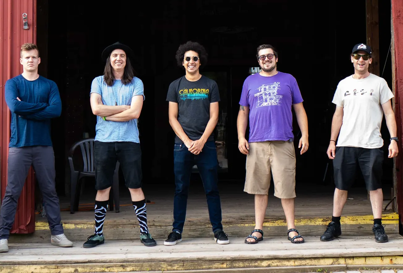 Someone standing on a wooden porch of a building with five other individuals, all casually dressed, smiling and posing for a