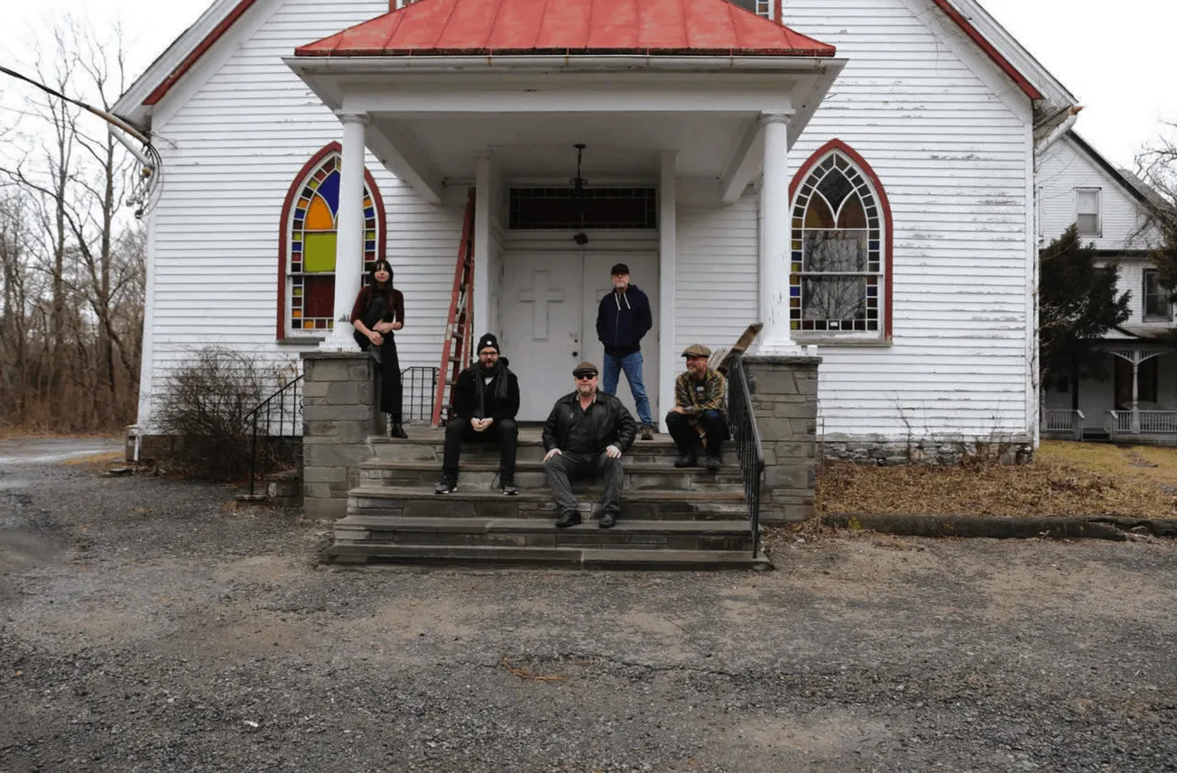 Abandoned white church with stained glass windows and six people sitting and standing on the front steps, creating a moody, a