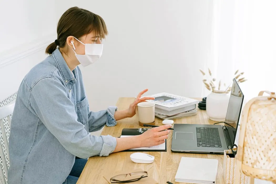 Woman wearing mask working on laptop at home with coffee, notebooks, and books on desk, modern workspace, remote work, digita