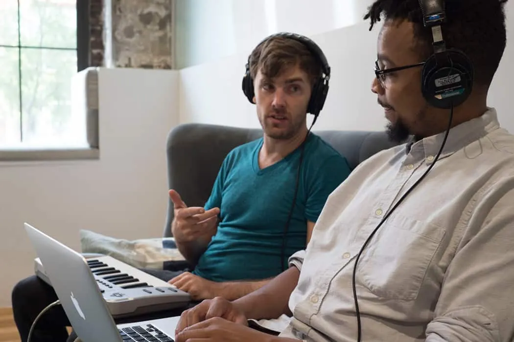 Young men collaborating on music production, using headphones and laptops, in a creative workspace.
