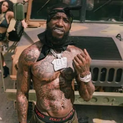 A shirtless man with tattoos, wearing jewelry and a bandana, standing in front of a Jeep during daytime outdoor event.