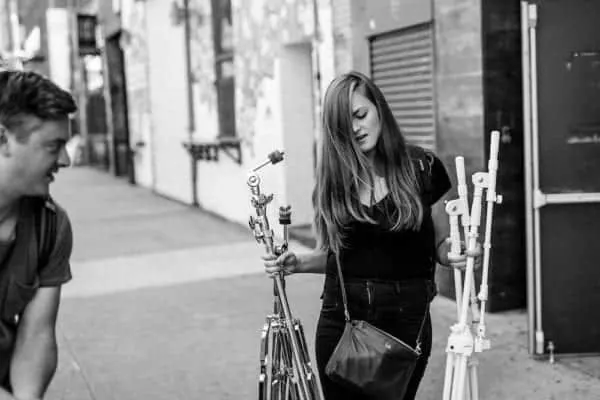 A young woman browsing street art supplies outdoors, holding spray paint cans and stencils, with a man observing nearby, show