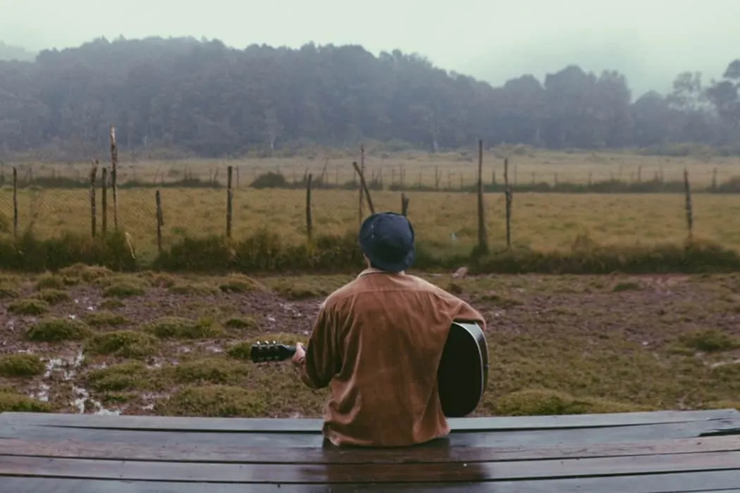 Cityscape, outdoor music performance, person playing acoustic guitar on wooden bench, nature setting with rolling hills, fenc