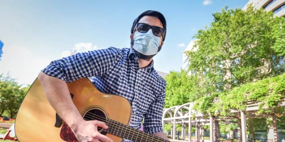 Percussionist, playing acoustic guitar outdoors, wearing sunglasses and face mask, vibrant park scene with green trees and bl