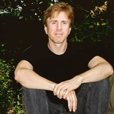 A man sitting outdoors in a nature setting, casually posing with a relaxed expression, wearing a black t-shirt and jeans.