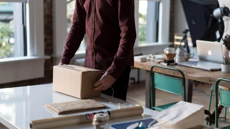 A person preparing a package on a desk in a modern, well-lit office space with large windows, workspace essentials, and a cre