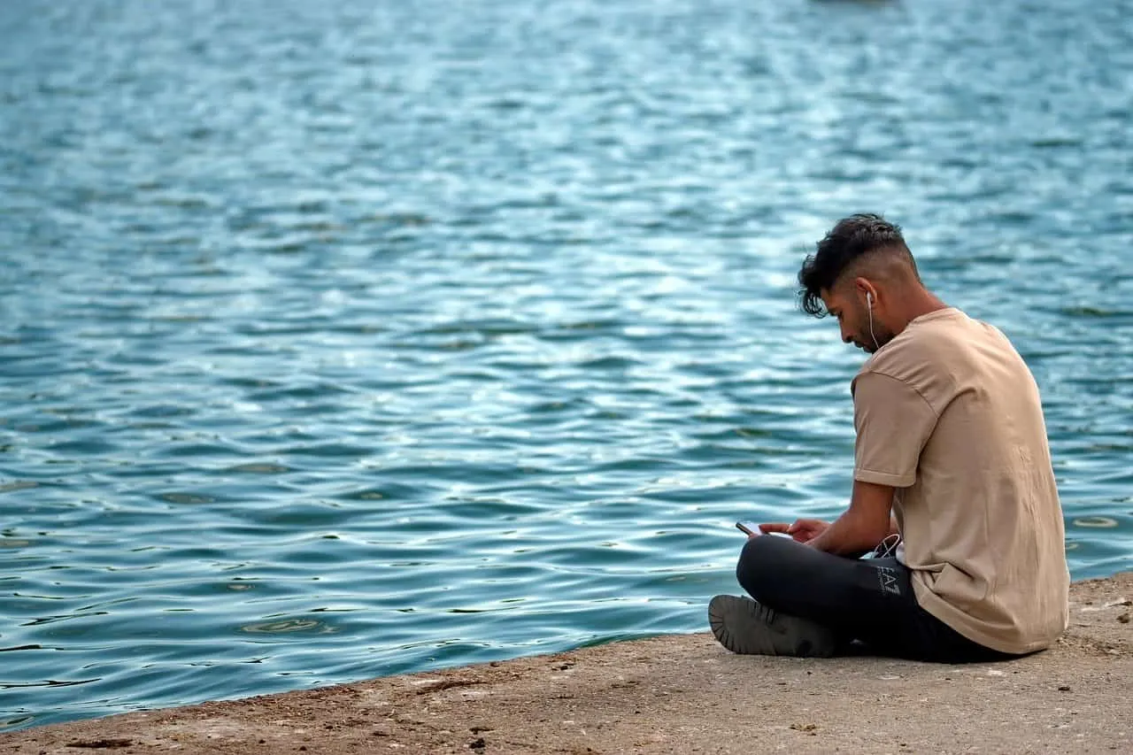Young man sitting by the water using a smartphone, listening to music with earphones, relaxing by the lakeside, with clear bl