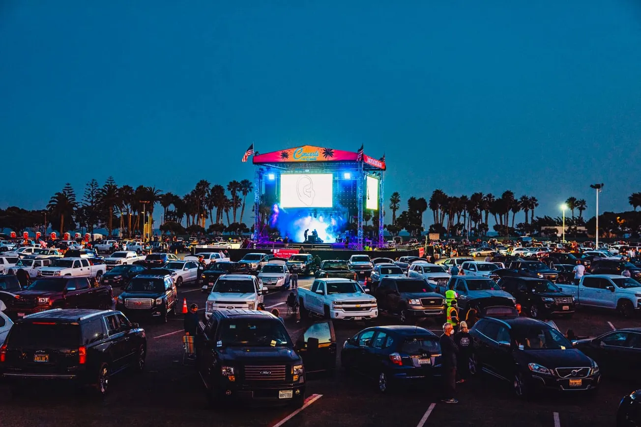 Concert event on a large outdoor screen at a drive-in music festival with a parking lot filled with cars, palm trees in the b