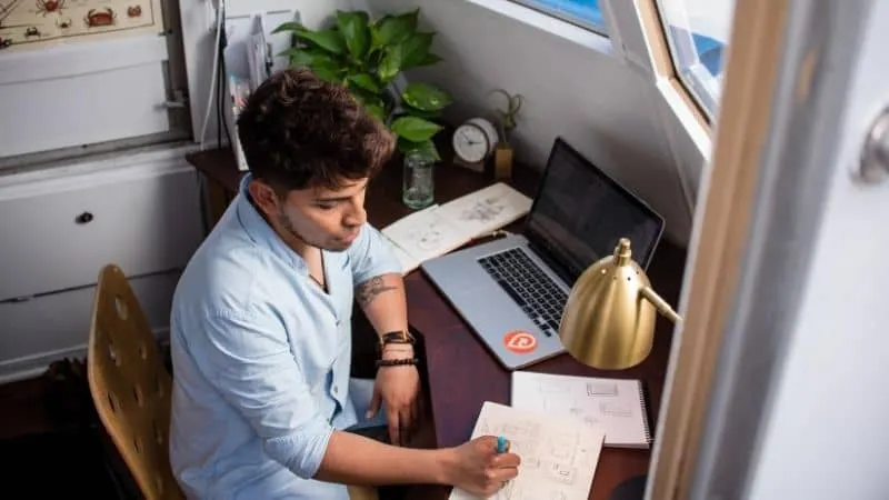 Cozy home workspace with a young man working on sketches at a wooden desk, surrounded by a laptop, green plant, and natural l