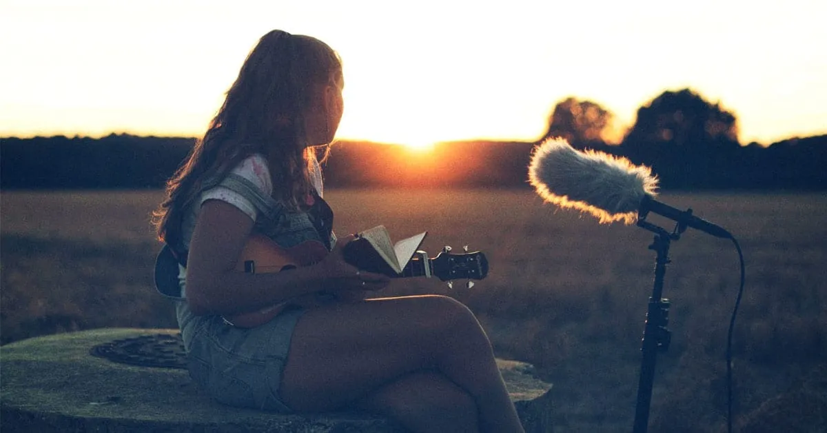 A young woman playing guitar outdoors at sunset, recording vocals with a professional microphone, creating music in a natural