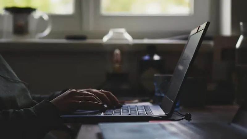 Person typing on a laptop in a dimly lit workspace, with natural light coming from the window. Focus on digital work, technol