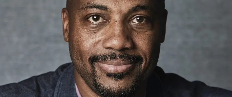 Celebrity portrait of a man with a beard and mustache, smiling, against a gray background.
