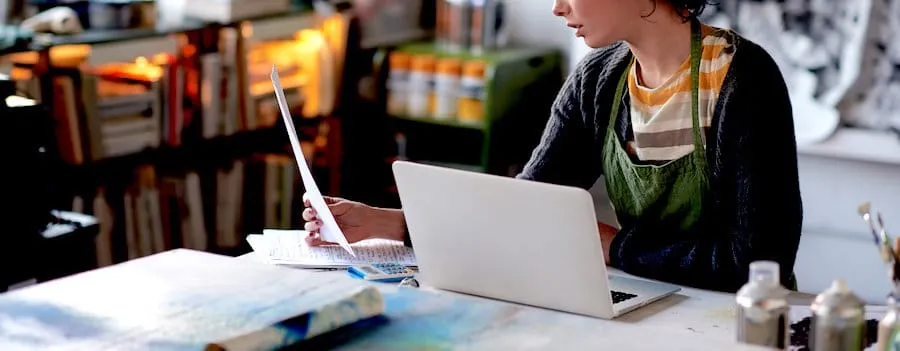 Young woman working on a creative project with a laptop and printed papers in a cozy, modern workspace.