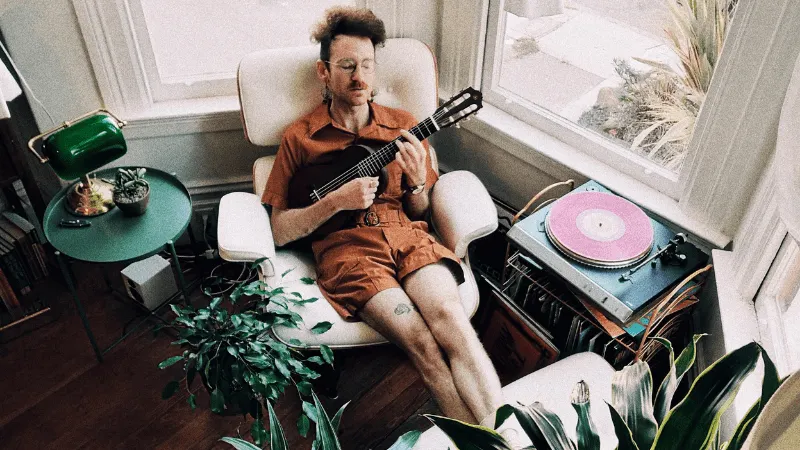 Man playing guitar in cozy home setting with vinyl turntable and green plants.