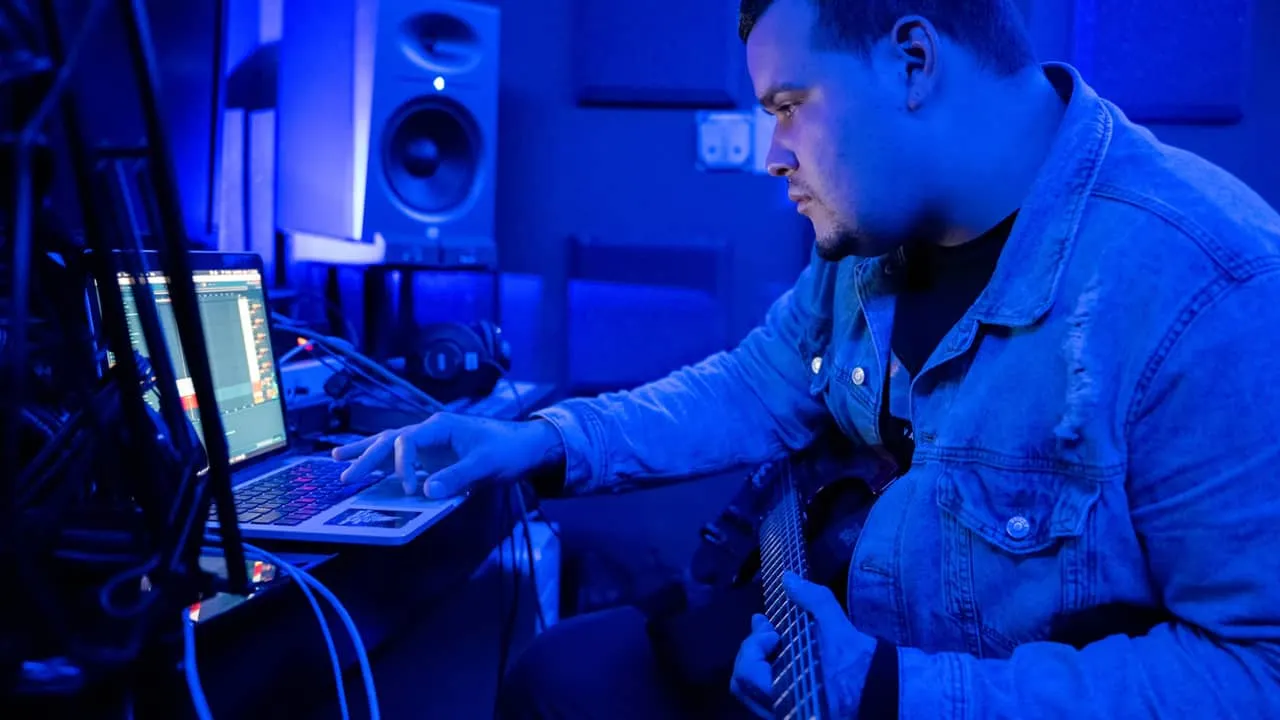 Music producer working on music production in a dark, blue-lit studio with a laptop and guitar.