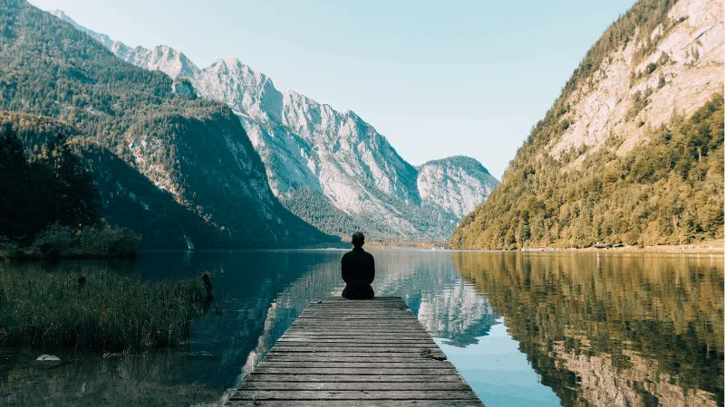 Serene person meditating on wooden dock by mountain lake, peaceful nature scene for mental wellness, relaxation, mindfulness,