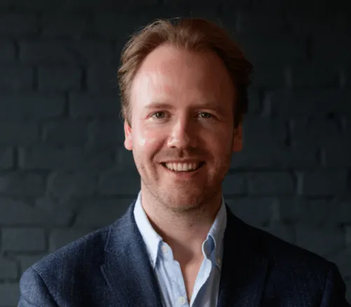 Bright professional portrait of a smiling man in business attire against a dark brick wall, representing leadership and exper