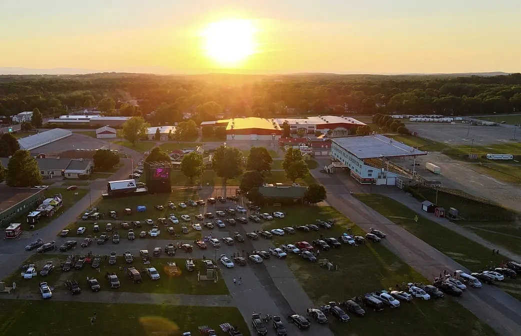 Sunset over outdoor concert venue with parked cars, stage, and surrounding buildings in a rural area, capturing live music ev
