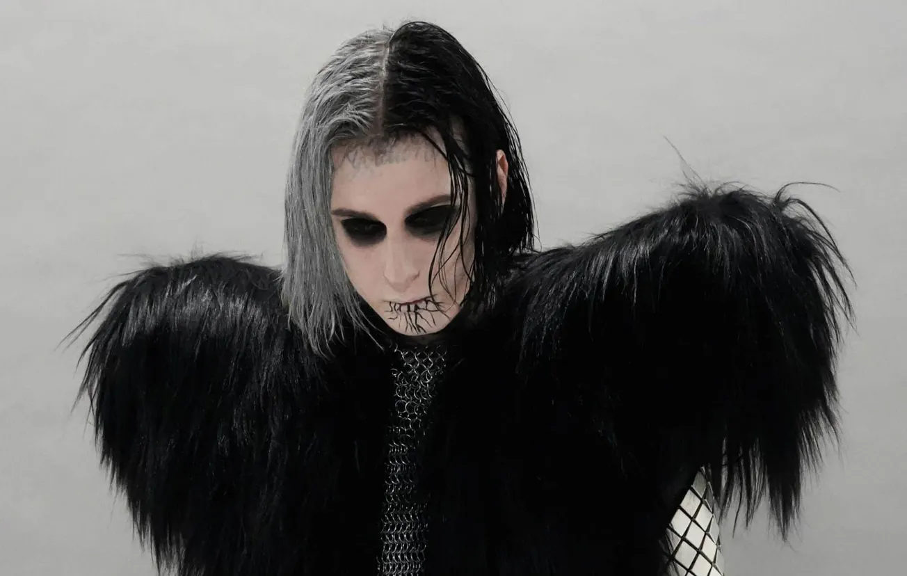 Dark goth musician with black makeup and dramatic hairstyle wearing black faux fur and fishnet clothing, embodying alternativ