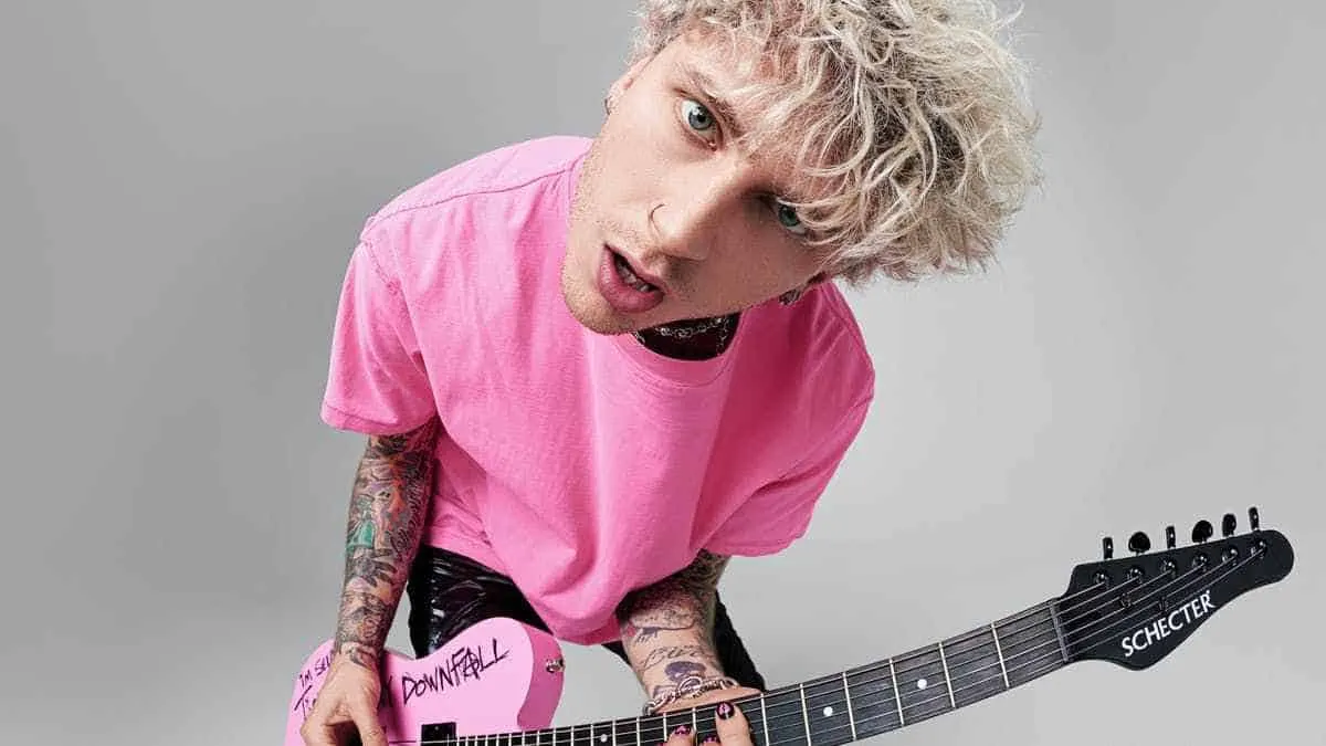 Viral musician with platinum blonde curly hair wearing a pink t-shirt playing a pink Schecter guitar against a plain gray bac