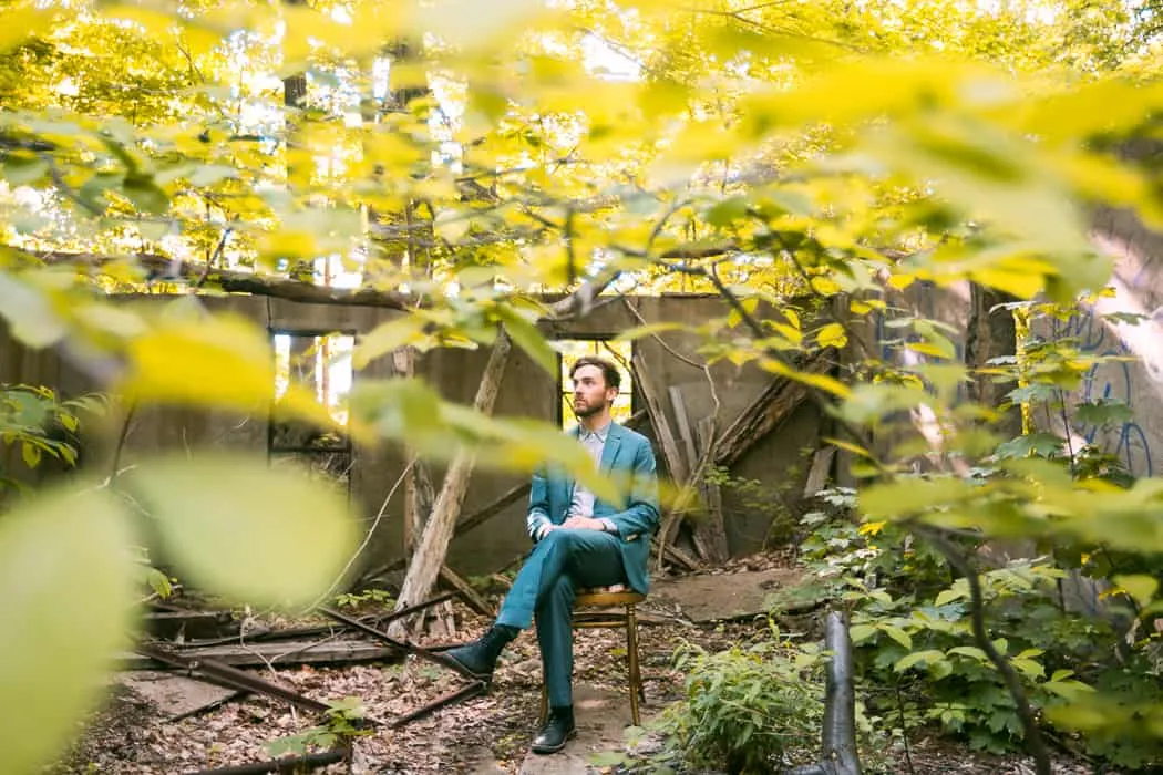 Abandoned urban outdoor space with a man in a suit sitting on a chair amid overgrown plants and graffiti-covered walls.
