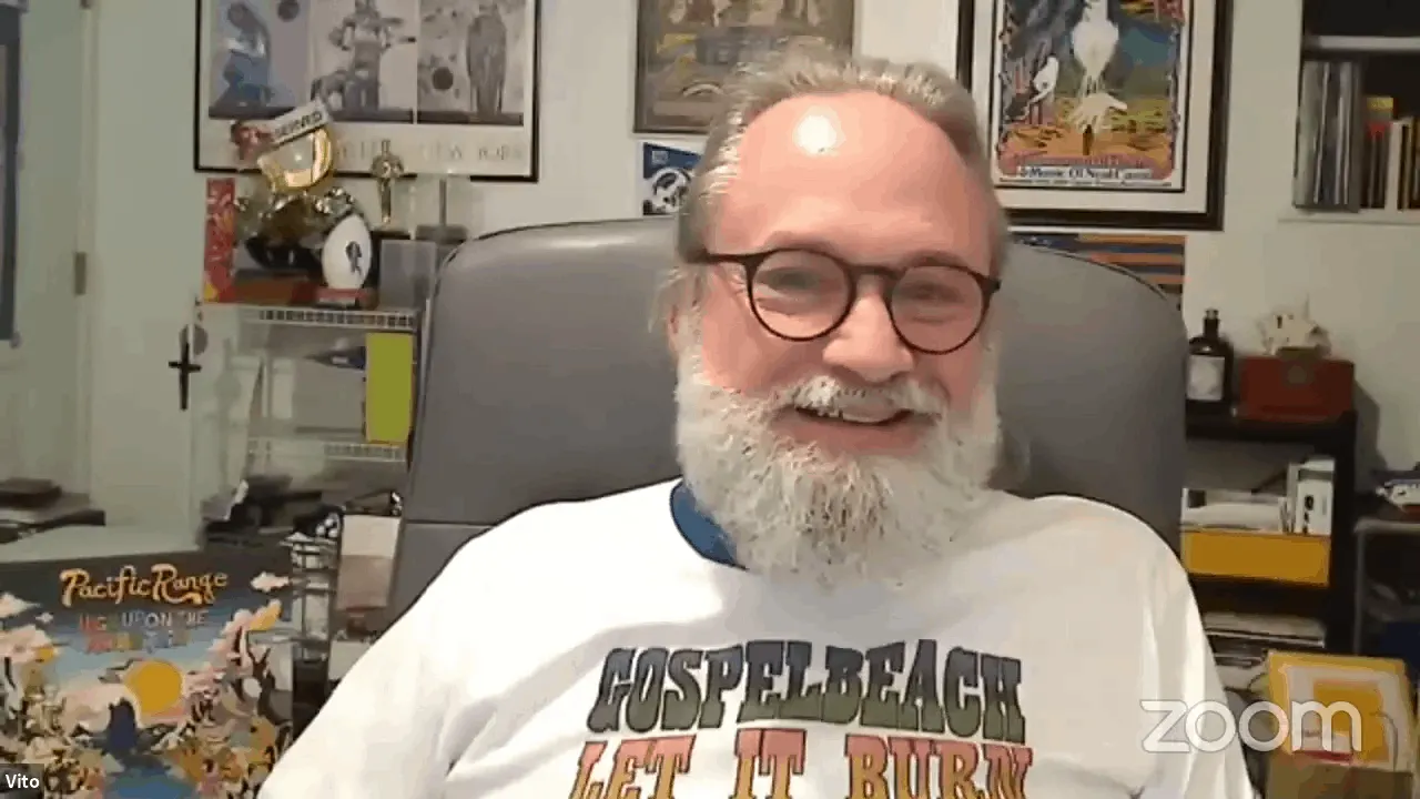 Smiling man with a beard and glasses sitting in an office, surrounded by colorful posters, books, and collectibles.