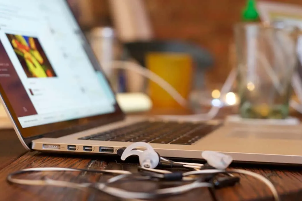 Laptop open with music streaming page on screen, white earphones resting on wooden desk, cozy studio environment, creative wo