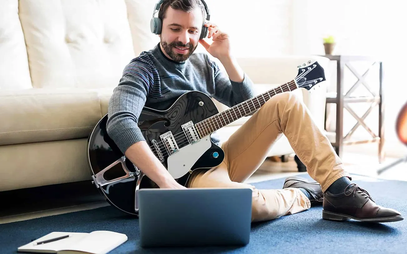 Guitarist wearing headphones with a laptop and notebook on the floor, enjoying playing music at home.